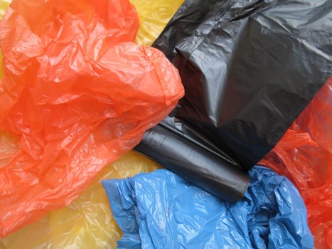 The image displays a close-up view of multiple crumpled plastic rubbish bags in various colors, including bright orange, black, blue, and yellow, scattered on a flat surface. The bags are made of thin, flexible plastic with shiny textures that reflect light. The orange bag is partially draped over a black bag, which appears to be filled or tied at the top, while the blue and yellow bags are lying flat, showing their crinkled surfaces. The background is not clearly visible but suggests an outdoor or utilitarian environment, likely related to waste collection or disposal activities. The scene is well-lit, highlighting the different textures and vibrant colors of the plastic bags, which are indicative of household or commercial waste temporarily stored or ready for collection by a rubbish removal service, such as Commercial Waste Lee, serving the Lee area near postcode [POSTCODE]. This visual offers a clear depiction of waste items commonly handled during rubbish clearance operations in local urban or suburban settings.