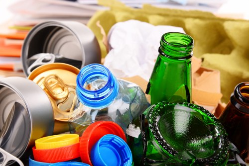 A close-up image capturing a collection of discarded glass and plastic bottles, including a prominently placed green glass bottle and a clear plastic bottle with a blue cap, both lying amidst various other plastic containers and bottle caps in shades of yellow, orange, and blue. Some bottles are broken or crumpled, with a crumpled piece of white paper and yellow packaging material visible in the background. The items are scattered on a surface that appears to be part of an indoor or outdoor area used for rubbish collection or disposal, with the surrounding environment showing no specific defining features. The scene reflects typical waste materials found during rubbish removal services, consistent with operations in Lee, and is managed by Commercial Waste Lee, supporting local waste management needs.