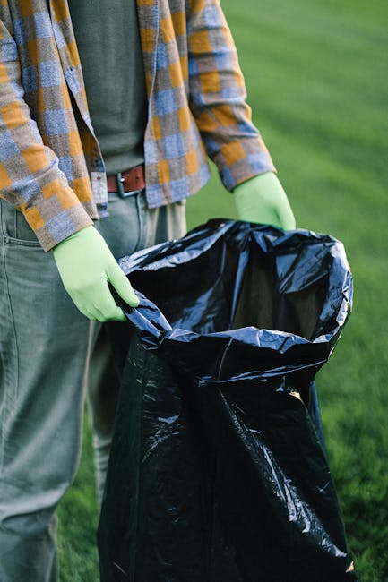 A person wearing a checkered yellow and gray shirt, green gloves, and gray trousers is holding open a large black plastic rubbish bag outdoors on a grassy area. The bag is partially filled and appears
